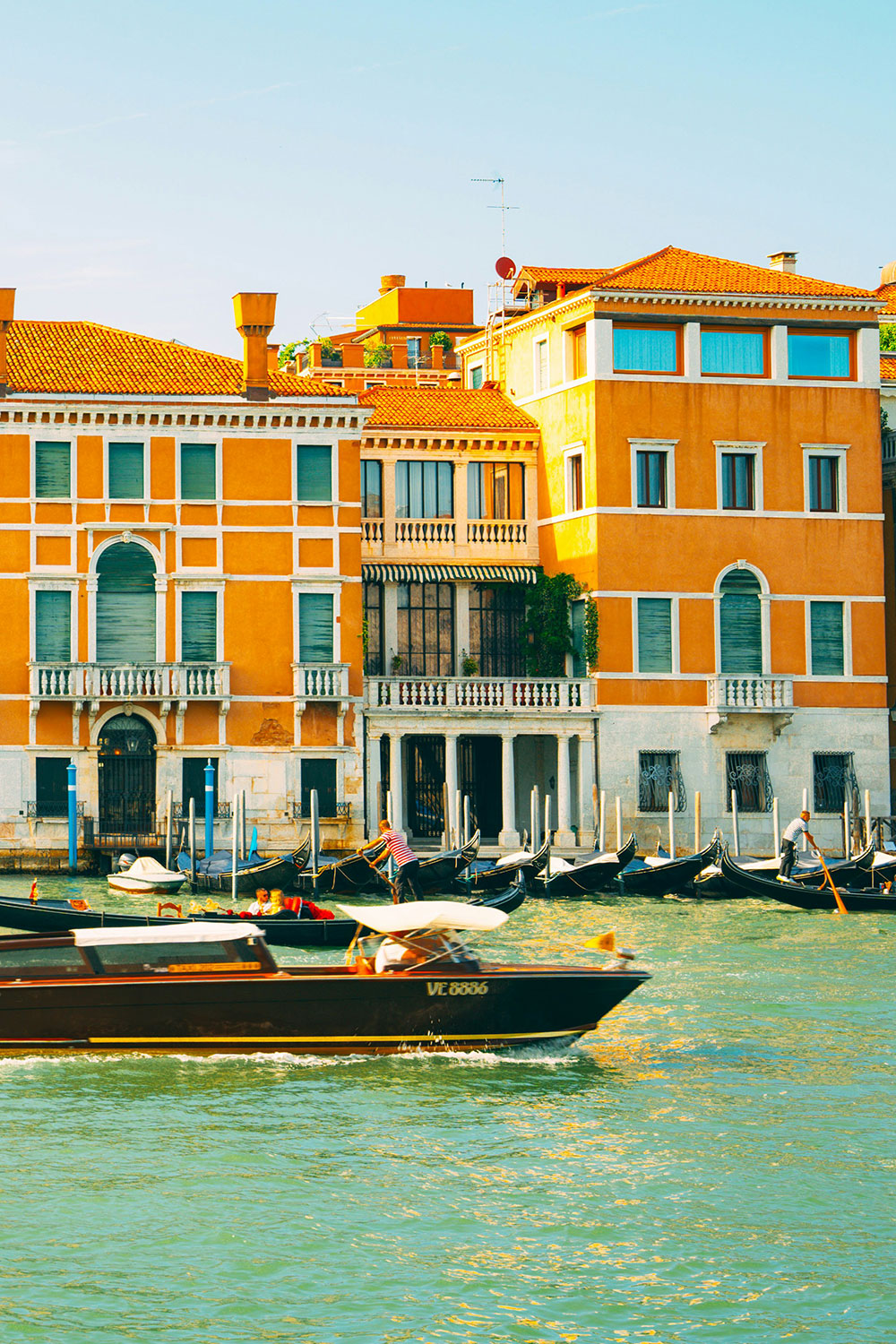 paesaggio veneziano, vediamo una barca scura che sta procedendo nel canale, nello sfondo vediamo dei palazzi arancioni