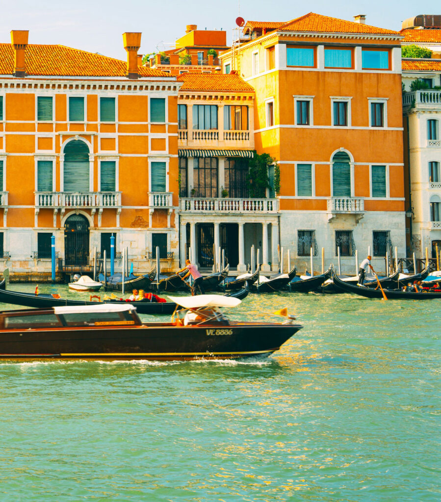 paesaggio veneziano, vediamo una barca scura che sta procedendo nel canale, nello sfondo vediamo dei palazzi arancioni